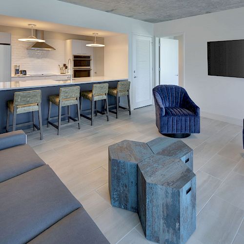 A modern open-concept living area with a kitchen, bar stools, a fridge, a TV, a blue sofa, two blue armchairs, and two wooden coffee tables.