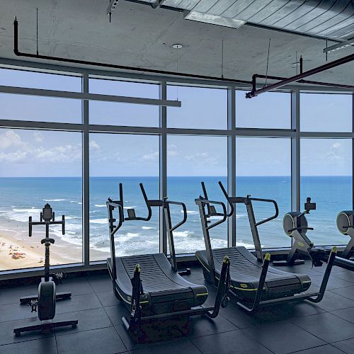 A gym with exercise equipment overlooks a scenic beach and ocean view through large floor-to-ceiling windows, offering an inspiring workout environment.