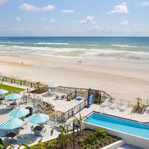 A beachfront scene with lounge chairs, sun umbrellas, a pool, a sandy shore, and ocean waves under a blue sky with scattered clouds.