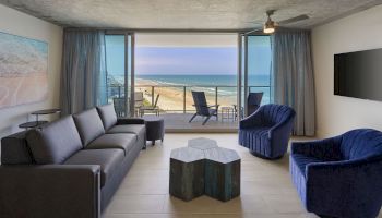 A modern living room with a beach view, large windows, black sofa, two blue chairs, a unique central table, and a wall-mounted TV.