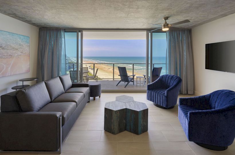 A modern living room with a beach view, large windows, black sofa, two blue chairs, a unique central table, and a wall-mounted TV.