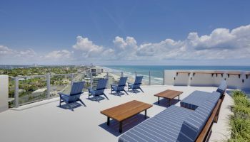 A rooftop deck overlooks the ocean, featuring blue cushioned lounge chairs, wooden tables, and a long cushioned bench under a clear blue sky.