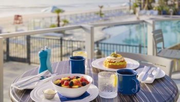 A breakfast spread including fruit salad, a pastry, coffee, and water on a table by a pool with a view of the beach in the background.