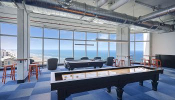 A spacious room with modern industrial design, featuring game tables, bar stools, large windows with an ocean view, and exposed ductwork on the ceiling.