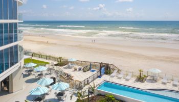 A beachfront scene with a modern building, outdoor pool, lounge chairs, and umbrellas overlooking a sandy beach and ocean under a sunny sky, ending the sentence.