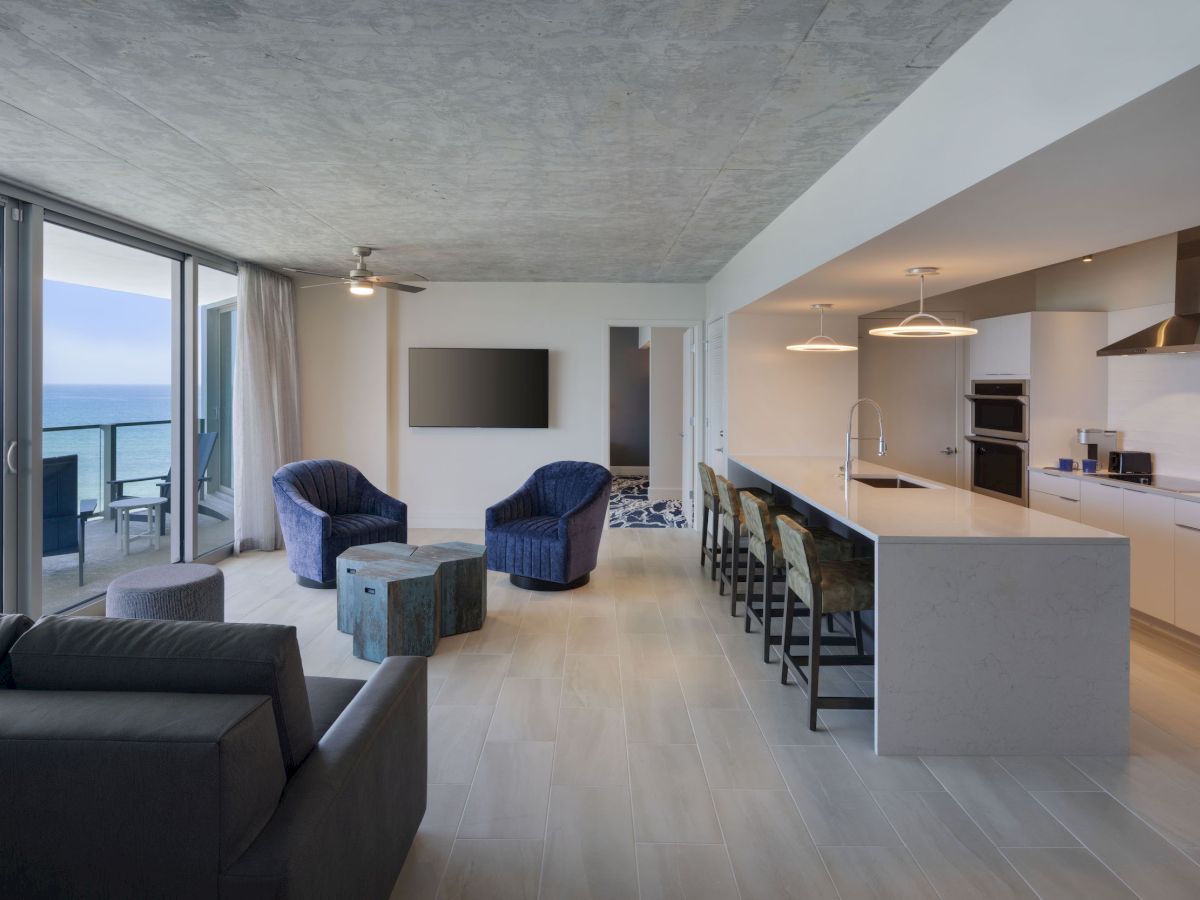 A modern living room and kitchen with ocean view, featuring a gray couch, blue chairs, a sleek kitchen island, and a balcony.