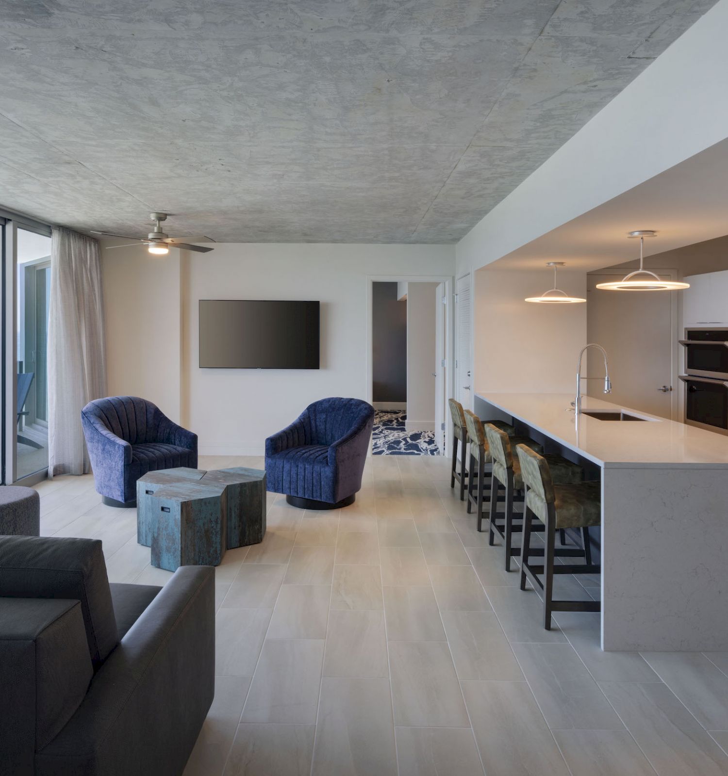 A modern living room and kitchen with ocean view, featuring a gray couch, blue chairs, a sleek kitchen island, and a balcony.