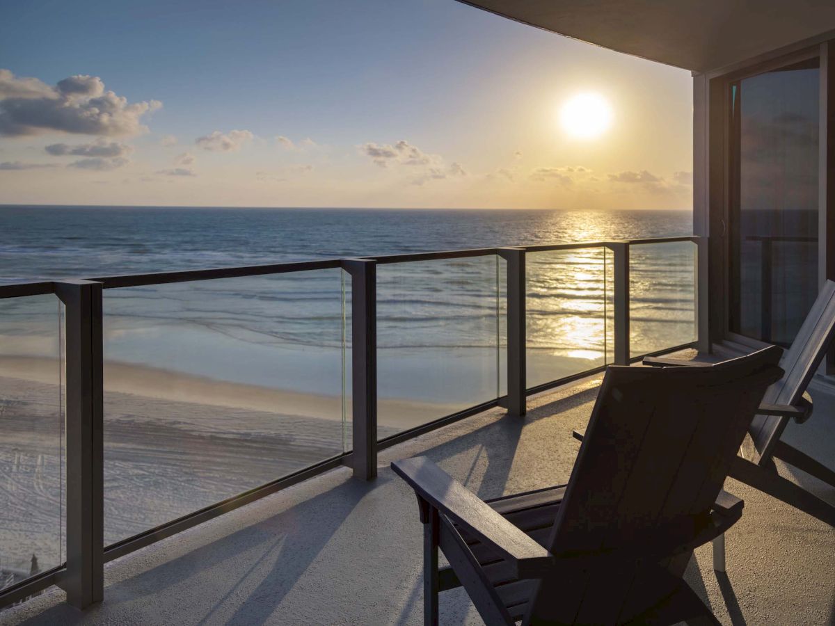 A balcony with two chairs overlooks a serene beach and ocean with the sun setting on the horizon, casting a warm glow.