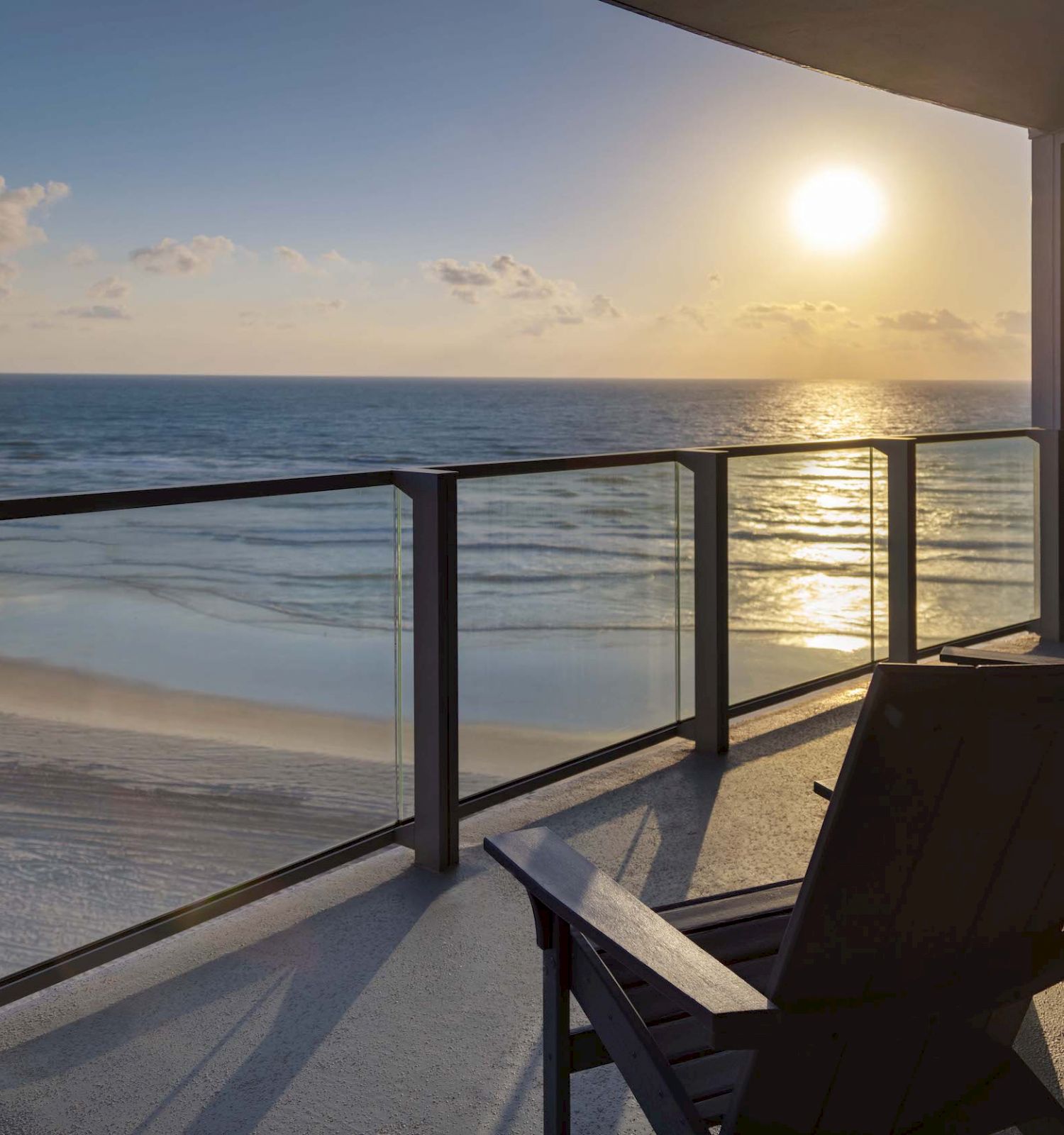 A balcony with two chairs overlooks a serene beach and ocean with the sun setting on the horizon, casting a warm glow.