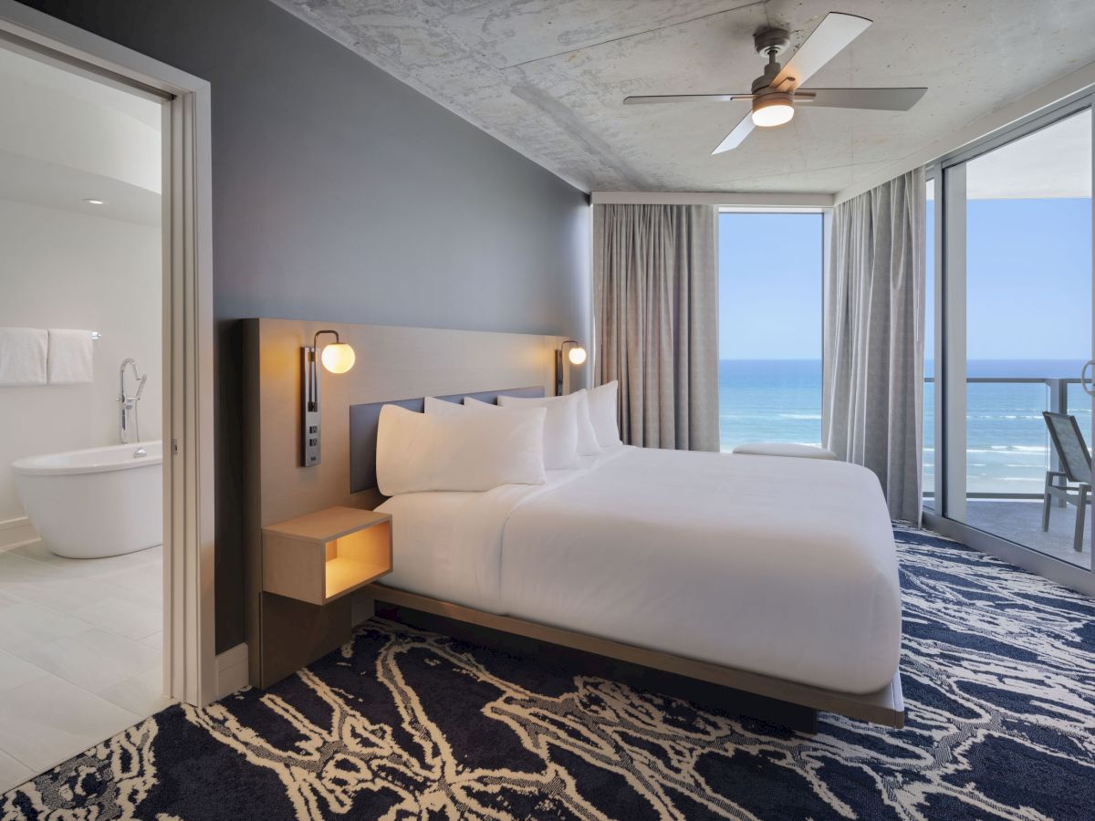 A modern hotel room with a large bed, ocean view balcony, stylish rug, and an adjacent bathroom featuring a freestanding tub ends the sentence.