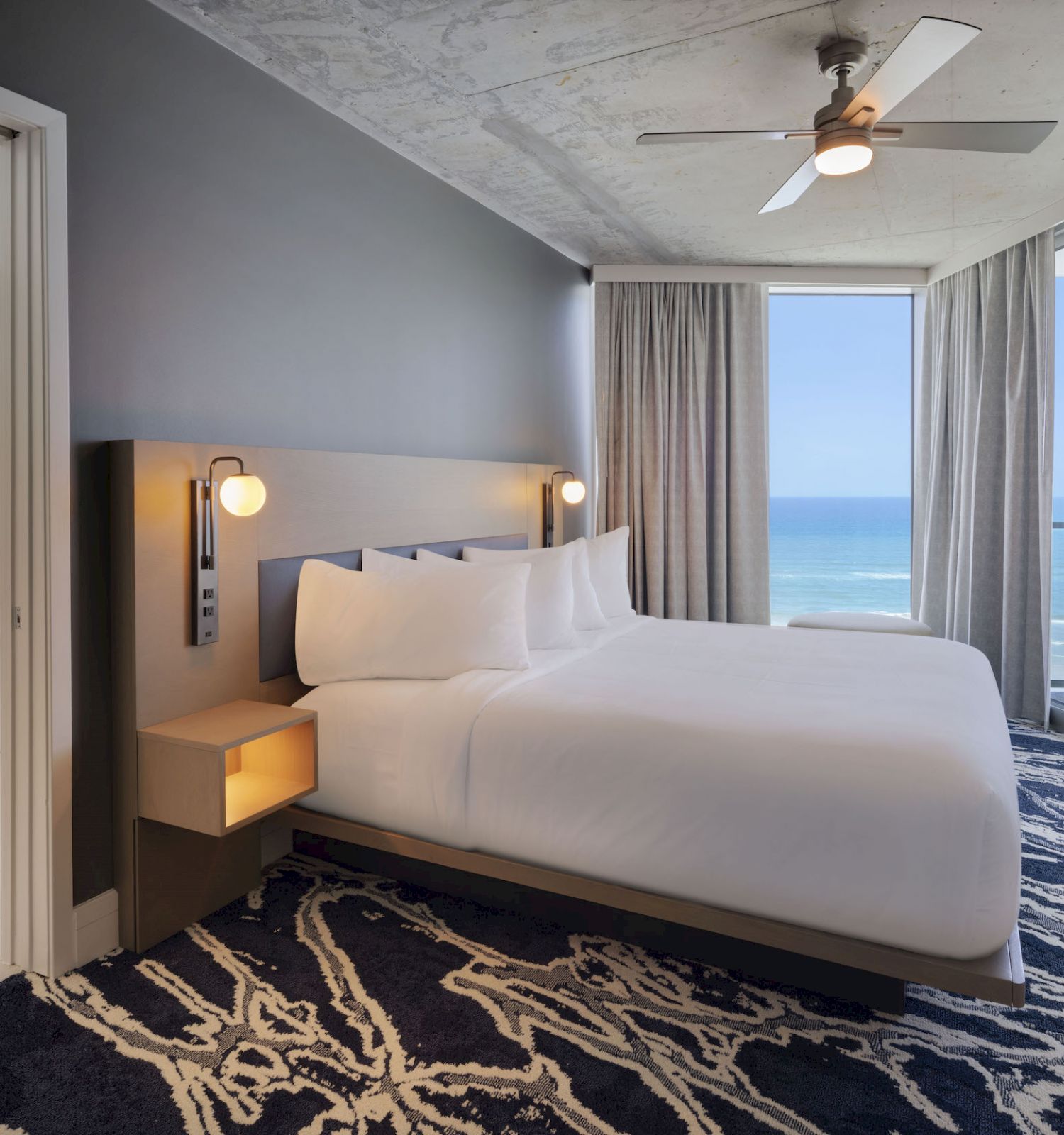 A modern hotel room with a large bed, ocean view balcony, stylish rug, and an adjacent bathroom featuring a freestanding tub ends the sentence.