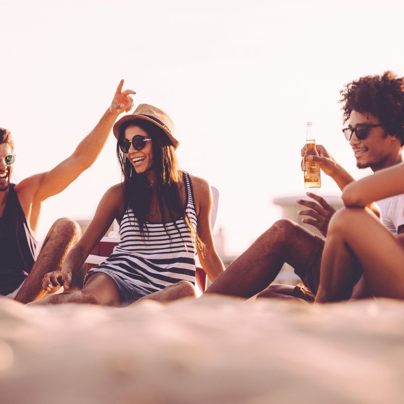 A group of four people sit on a beach, enjoying drinks and laughing together, with a sunny, relaxed atmosphere in the background.