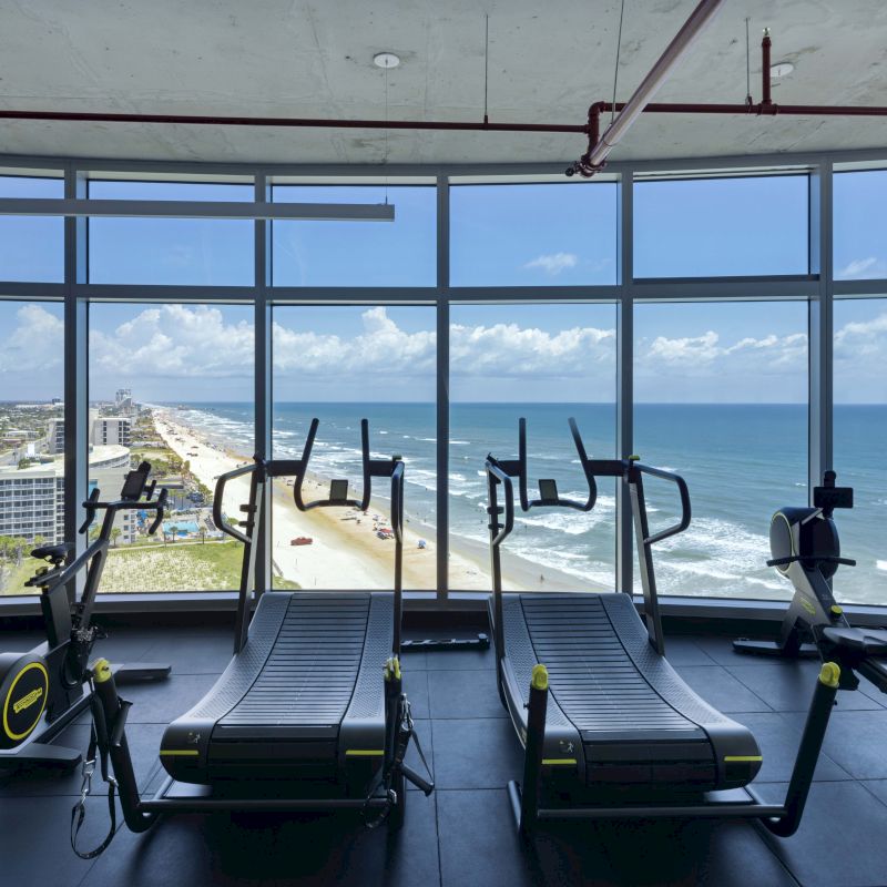 A gym with treadmills and exercise bikes inside a room with a panoramic window view of a beachfront and ocean.