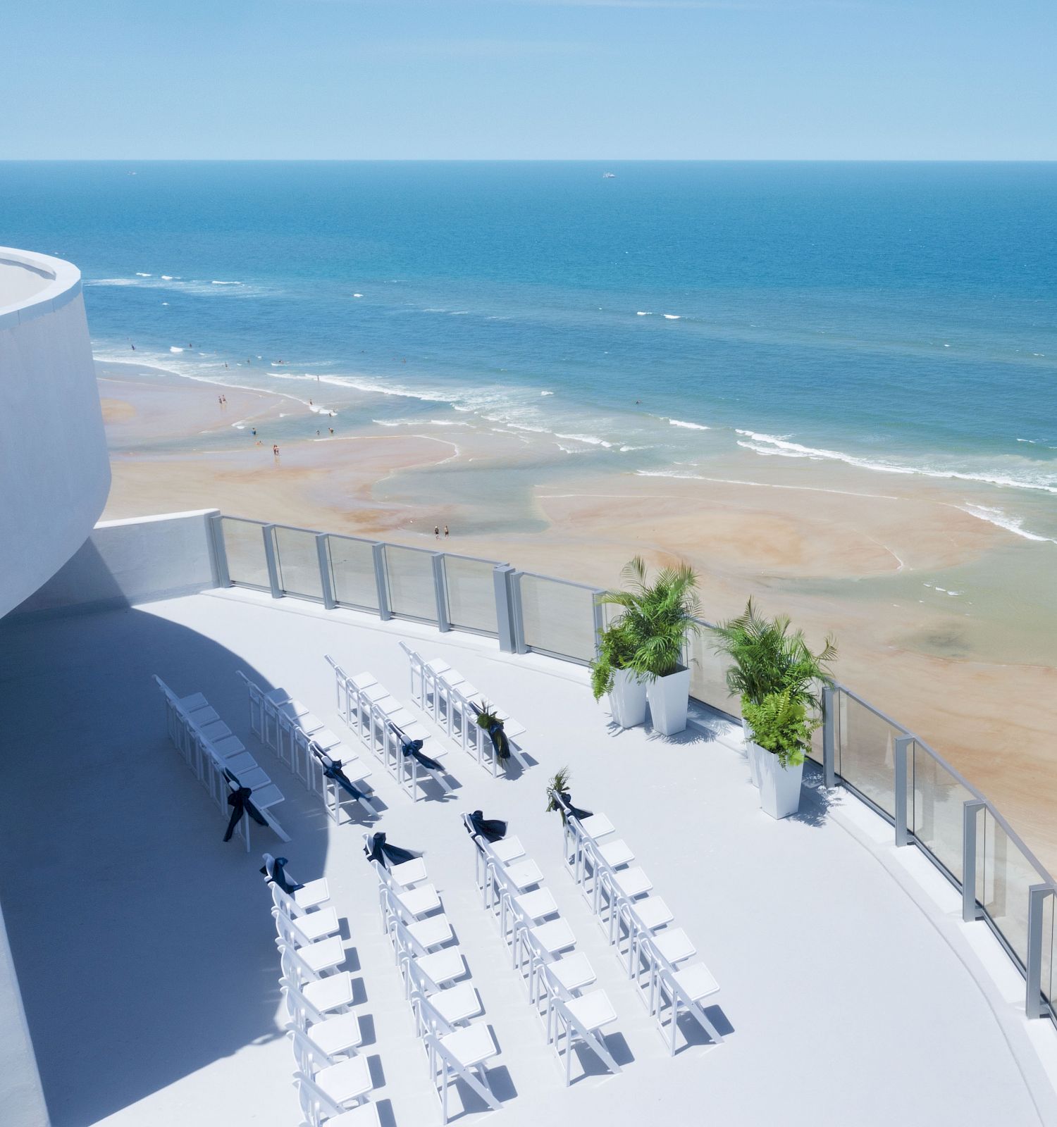 A modern seaside venue with white chairs arranged, facing the ocean. The scene is tranquil, with a few plants and a vast sandy beach below.