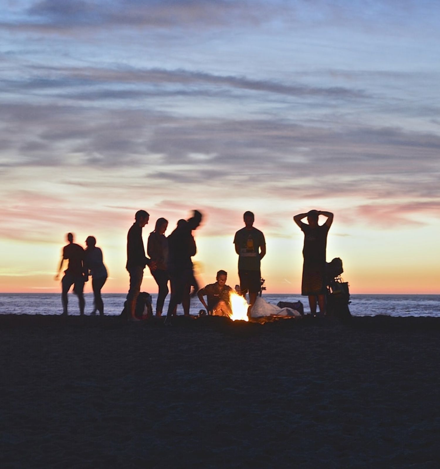 A group of people enjoying a beach bonfire at sunset, with the ocean and a colorful sky in the background.