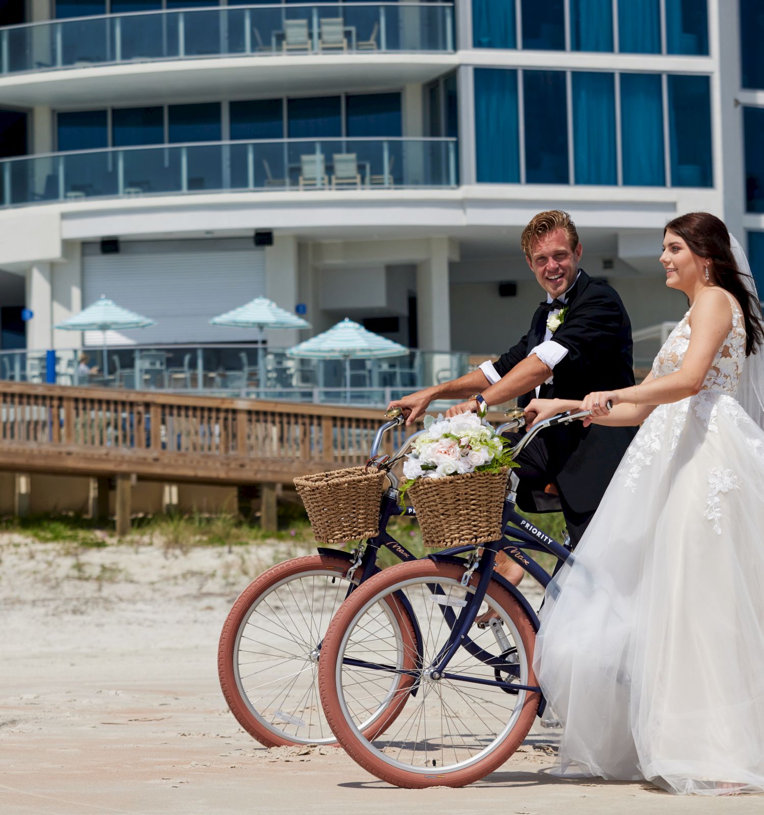 A couple in wedding attire rides bicycles on a beach, with a modern building and umbrellas in the background ending the sentence.